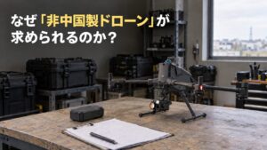 なぜ「非中国製ドローン」が求められるのか？ 「非中国製ドローン」を黄色で強調した題名と、作業台に置いた縦長の本体と吊り下げ式カメラを備えたドローンの写真風イメージ。